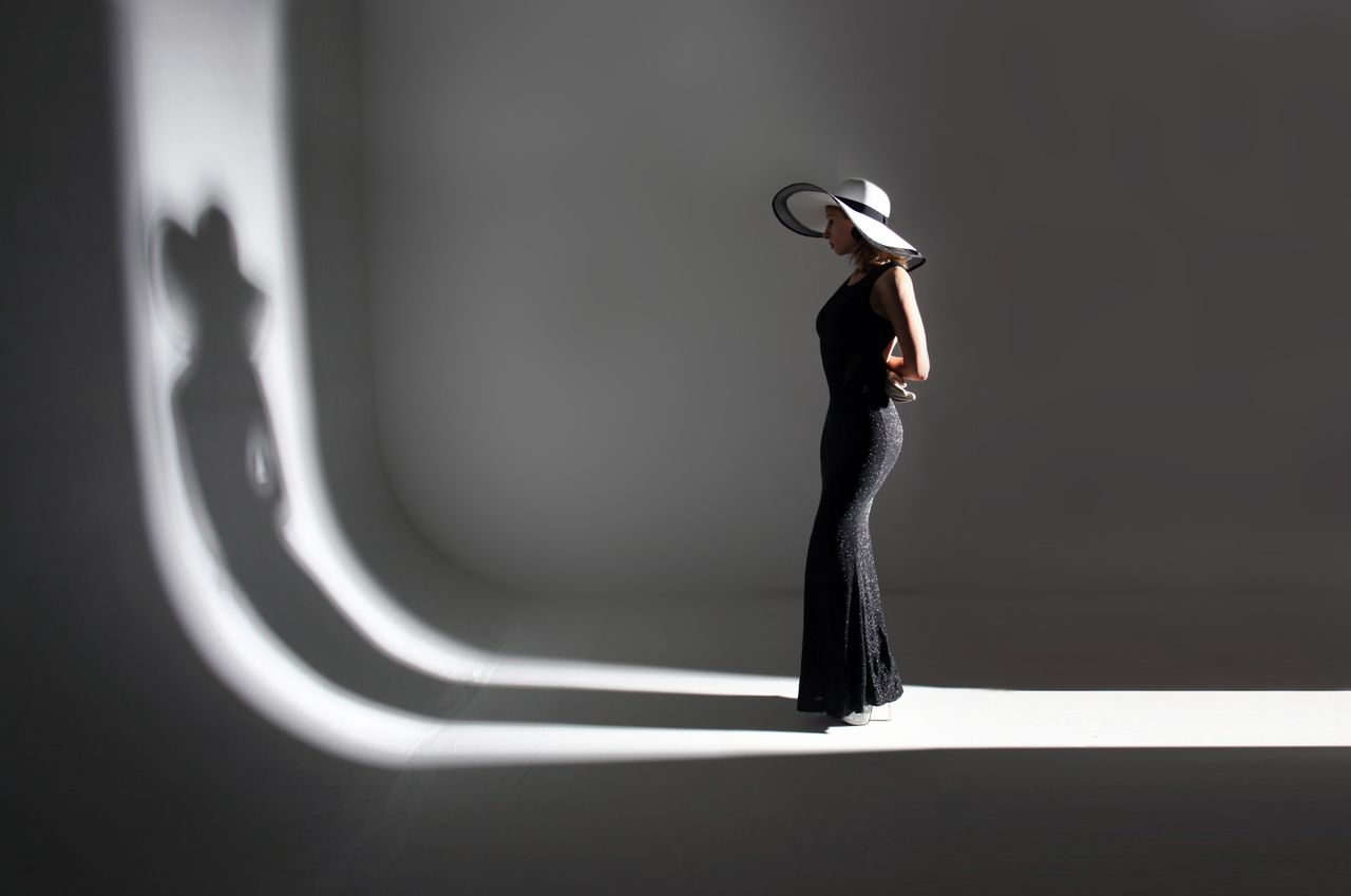 why-choose-us Full body of unrecognizable slim woman model in stylish long black dress and hat standing in dark studio and illuminated by light